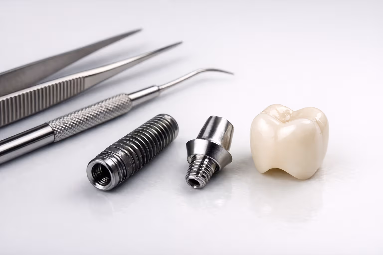 Close-up of a dental implant components including titanium post abutment and crown disassembled on a white sterile surface with dental instruments nearby