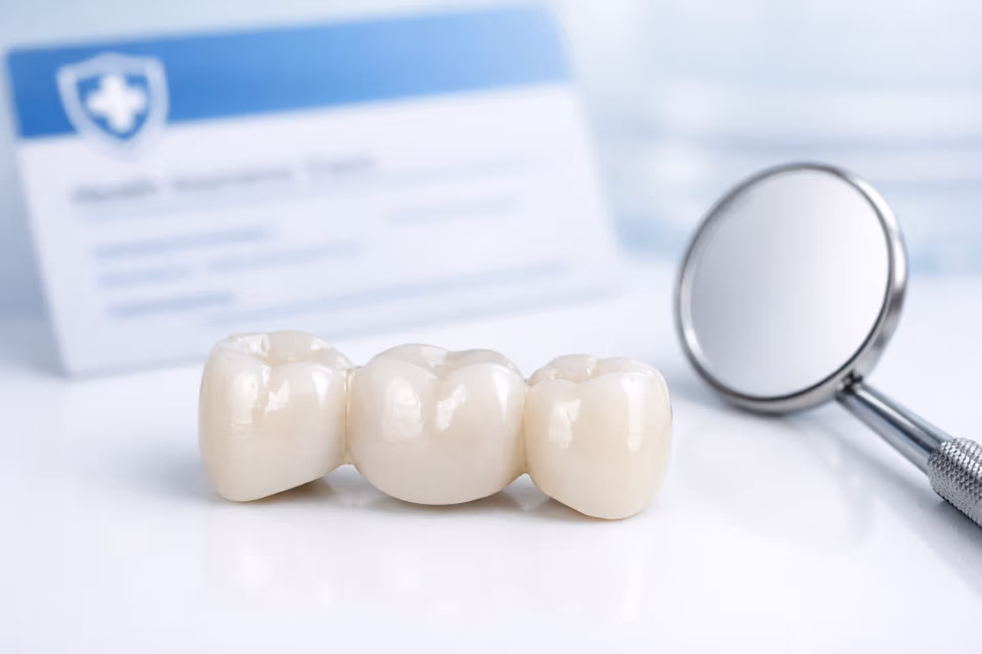 Three-unit dental bridge prosthetic on clean white surface next to dental mirror and blurred insurance card
