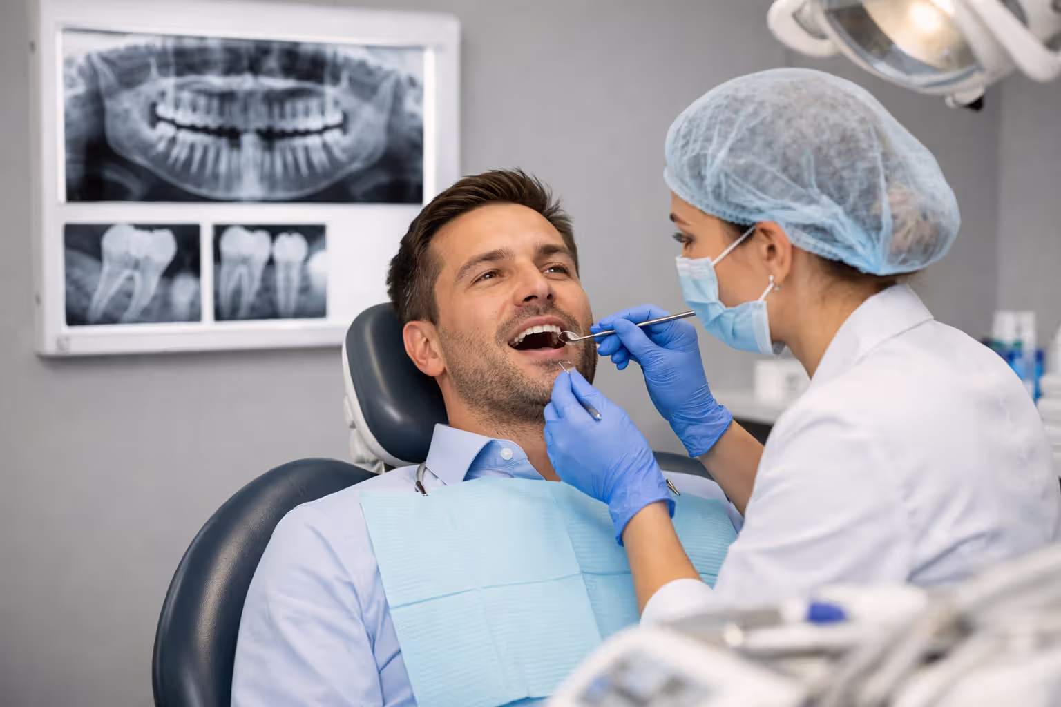 Adult man in a business shirt sitting in a dental chair while an orthodontist examines his teeth with a dental mirror, dental X-rays displayed on a light screen on the wall